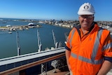 A man in a white hard hat and orange safety vest, stands on a high roof-top with Newcastle harbour in the background.