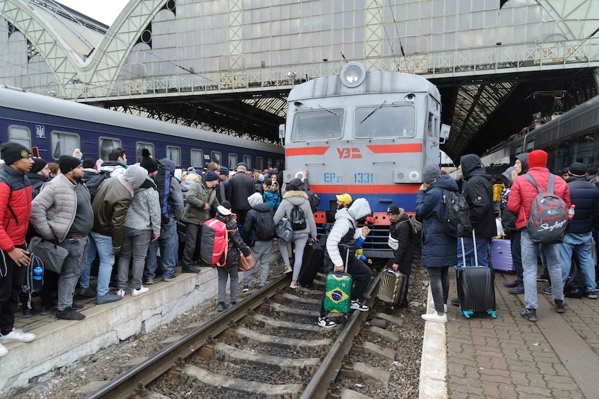 A crowd of people swarm a train, across the platform and across the tracks, trying to get on.