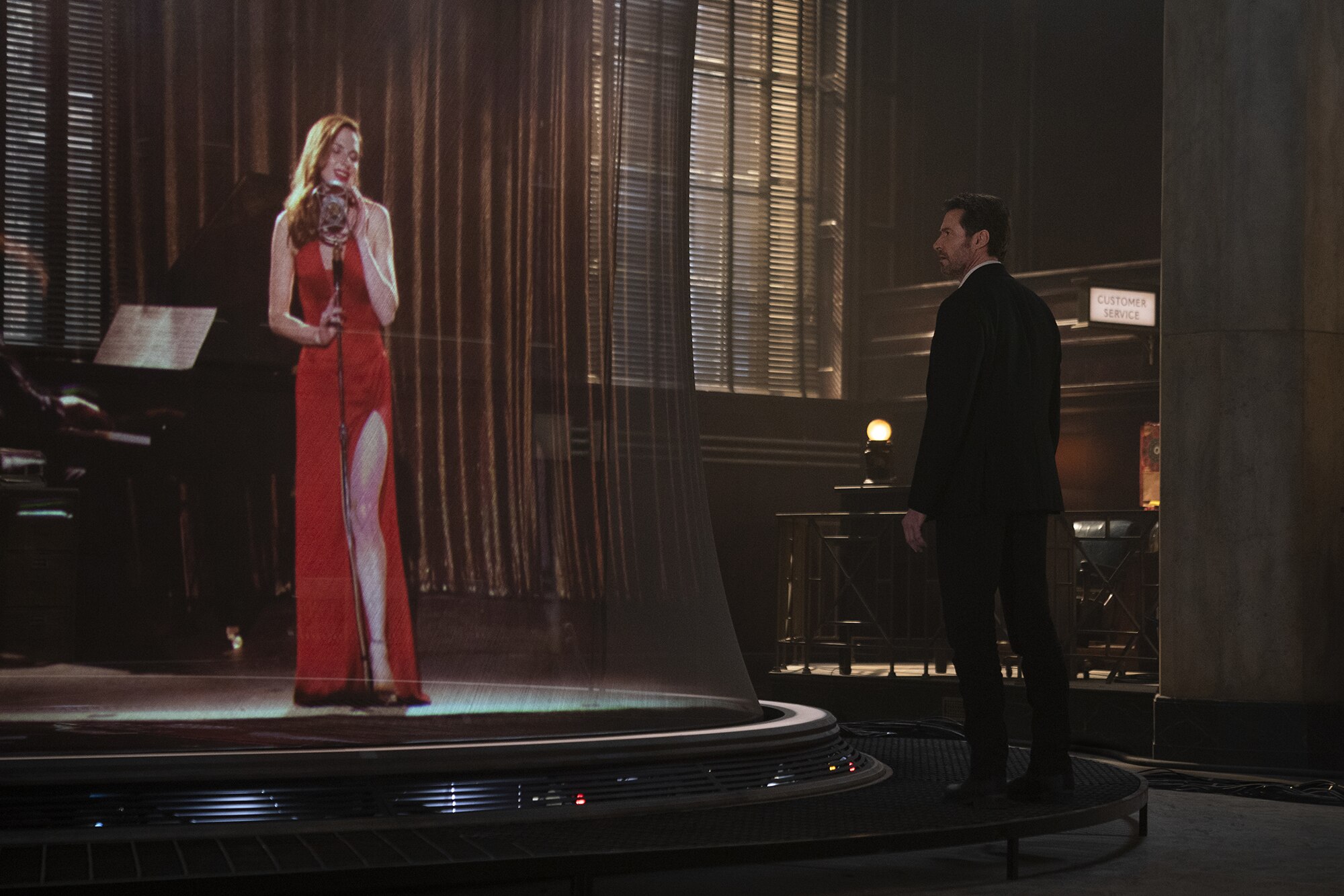 A holographic Rebecca Ferguson in a figure-hugging red dress sings at a microphone while Hugh Jackman looks on from shadows.