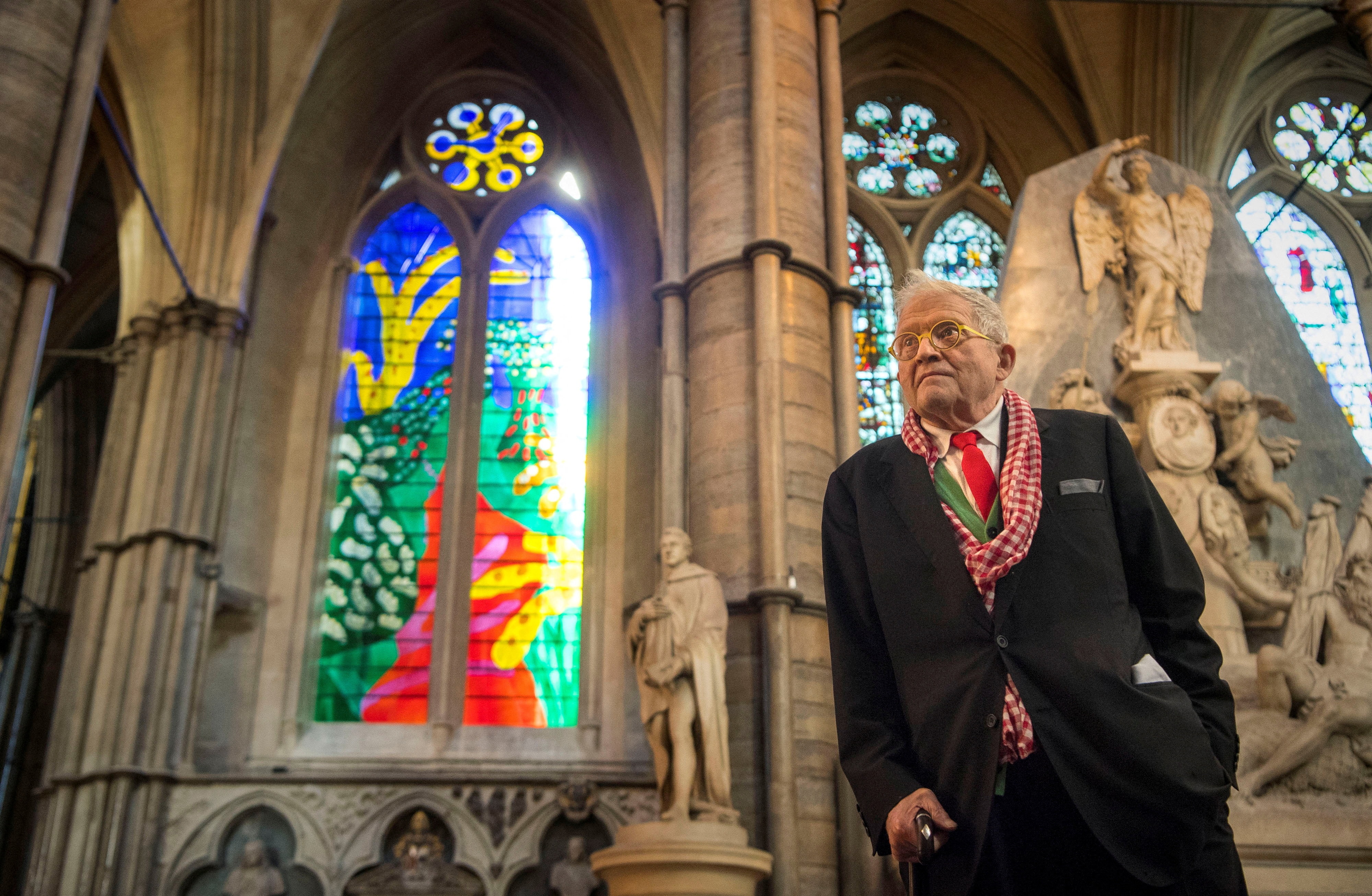 A silver haired artist looks thoughtfully and leans against wall in front of stained glass in church