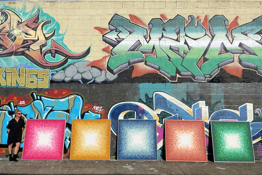 A woman stands in front of a graffitied wall, with artwork in the foreground.