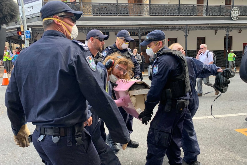 Male climate change protester with arm still in part of boat being led by 5 police officers.