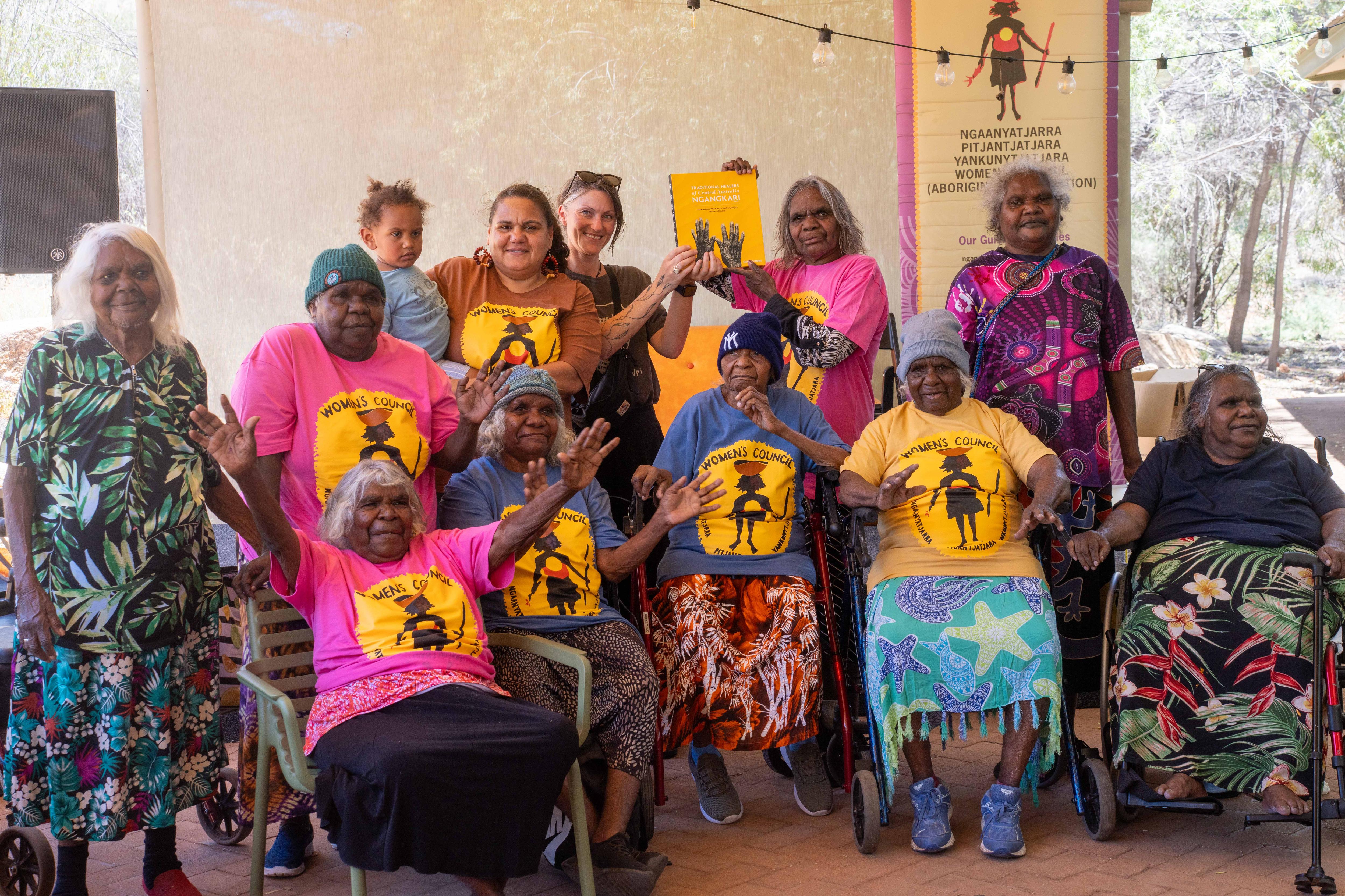 Un grupo de Ngangkari con camisetas del NPY posan para una fotografía grupal. Todos miran en una dirección diferente y hay manos en alto.