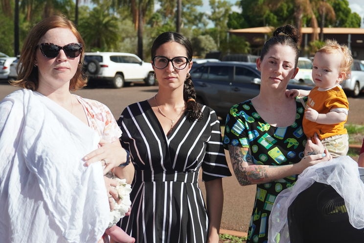 A group of women, one holding a baby, stand in a car park.