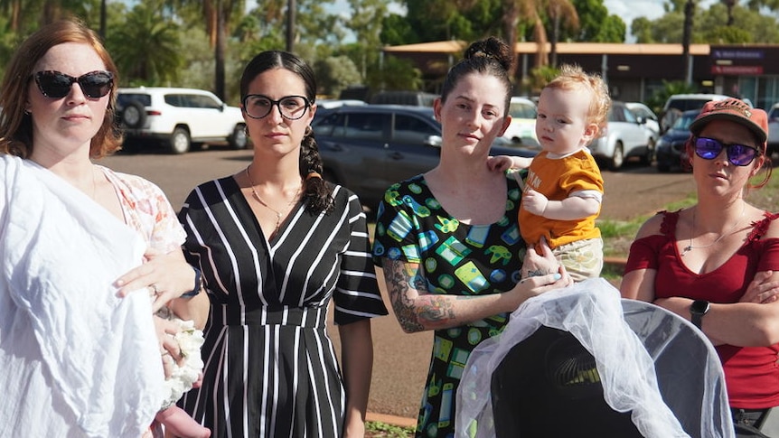 A group of women, one holding a baby, stand in a car park.