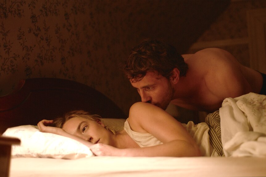A white man in his 30s kisses a white woman in her 30s in a bed; she looks away forlornly.