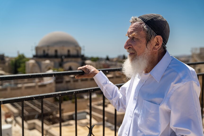 A man in a white shirt and black yarmulke with a long bushy beard looks out over a railing at Jerusalem