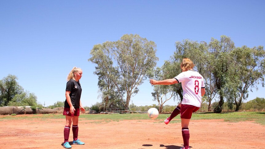 A young girls smiles at the camera, while in the background, her sister kicks a ball