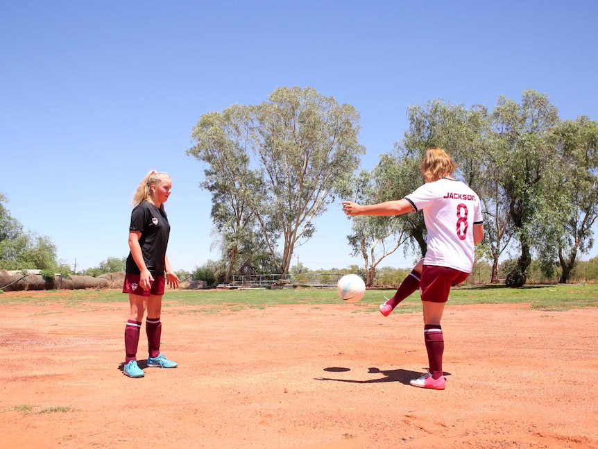 A young girls smiles at the camera, while in the background, her sister kicks a ball