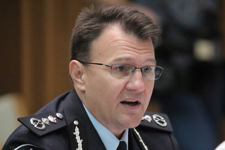 A man in a police uniform speaks into a microphone while seated at a desk next to a woman.