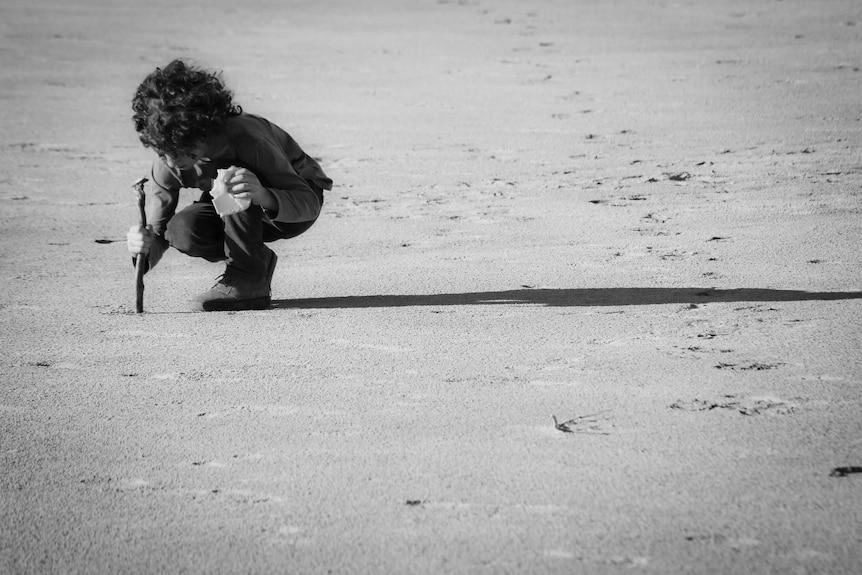A young boy draws pictures in the sand with a stick.