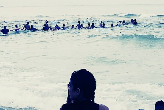 Group of people waist-deep in surf link arms in a line heading out to sea