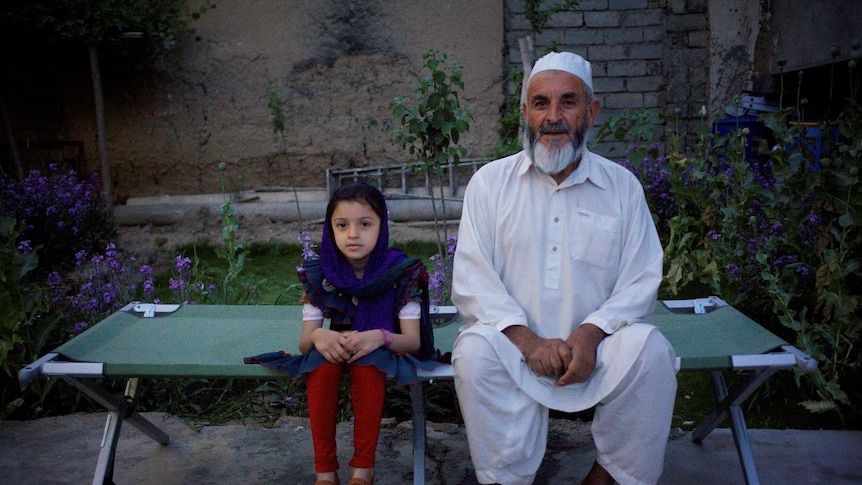 A man sits with his granddaughter on a military cot bed in his hospital garden in Helmand.