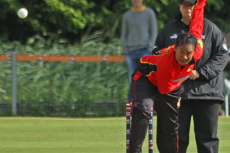 A PNG women's cricketer in a red and black top finishes her post-delivery stride as she sends down a ball. 