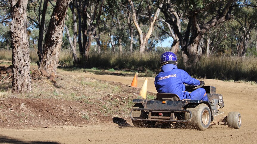 A man in a blue race suit drives a ride on lawn mower around a corner. 