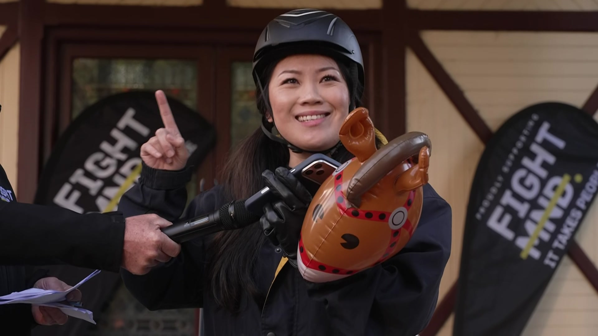 A woman wearing a black helmet holds a hobby horse in one hand and her finger up in air.