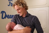 male midwife Christian Wright stands holding and looking at with three-week-old sleeping baby Imogen McQuire.