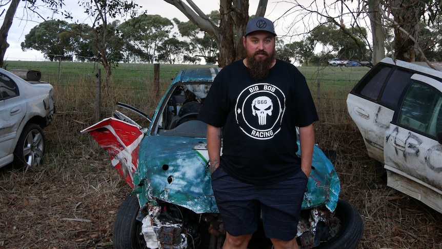 bearded man in t-shirt stands in front of destroyed car 