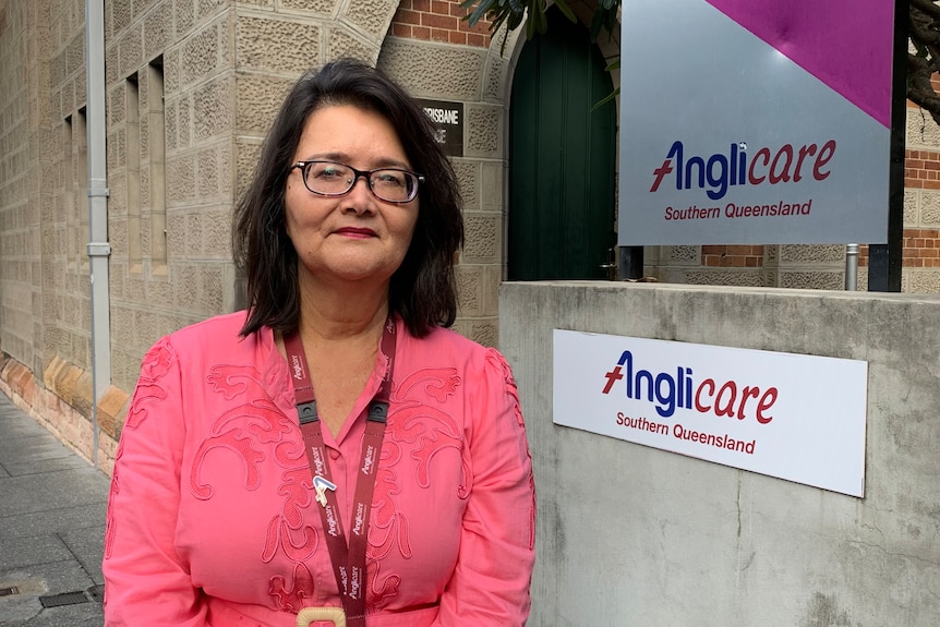 A woman in a intricate pink shirt posing next to an Anglicare sign on a church.