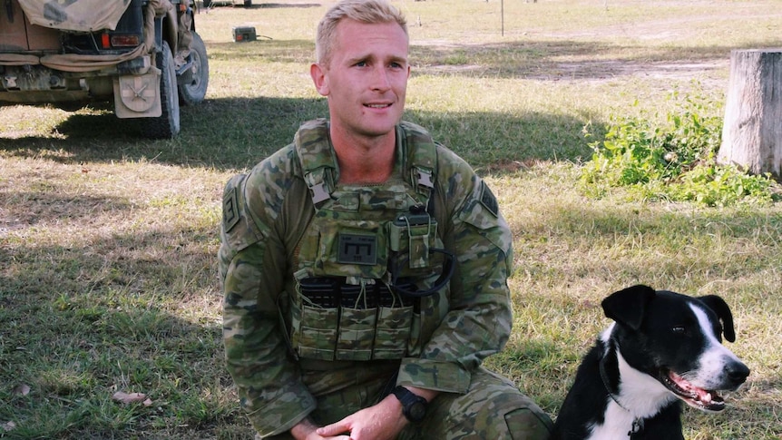 A man with blonde hair in an army uniform kneels next to a black and white dog