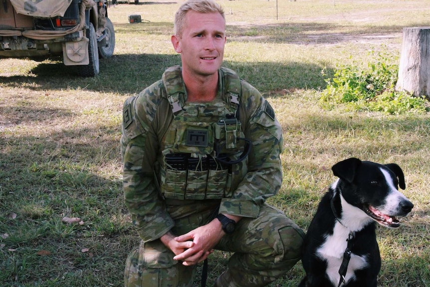 A man with blonde hair in an army uniform kneels next to a black and white dog