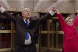 Alec Baldwin and Kate McKinnon stand holding their hands in the air at the entrance to the NBC studio building