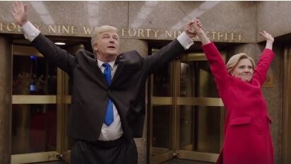 Alec Baldwin and Kate McKinnon stand holding their hands in the air at the entrance to the NBC studio building