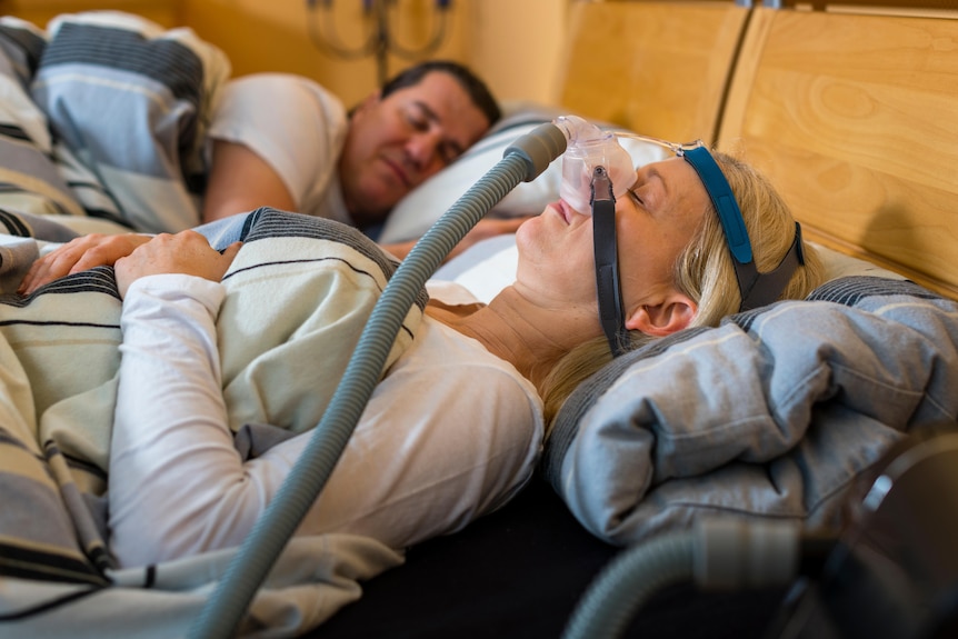 A woman is shown using a CPAP machine for obstructive sleep apnoea. Her male partner sleeps beside her.