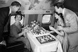 A black-and-white archival photo shows men and women in 1950s formal attire sitting on an aircraft eating from a luxury buffet