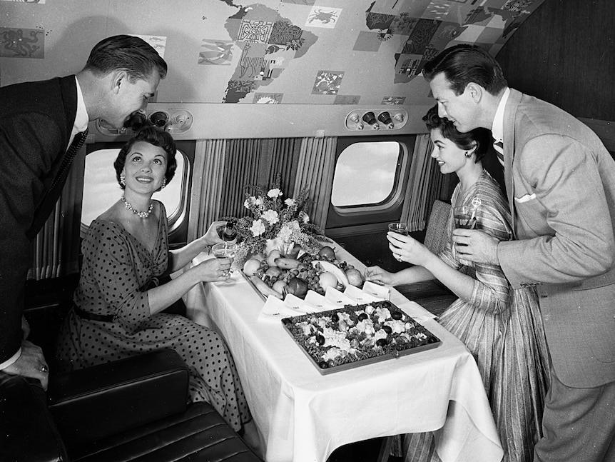 A black-and-white archival photo shows men and women in 1950s formal attire sitting on an aircraft eating from a luxury buffet