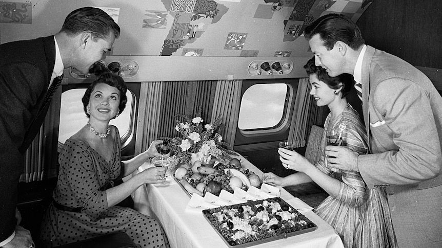 A black-and-white archival photo shows men and women in 1950s formal attire sitting on an aircraft eating from a luxury buffet