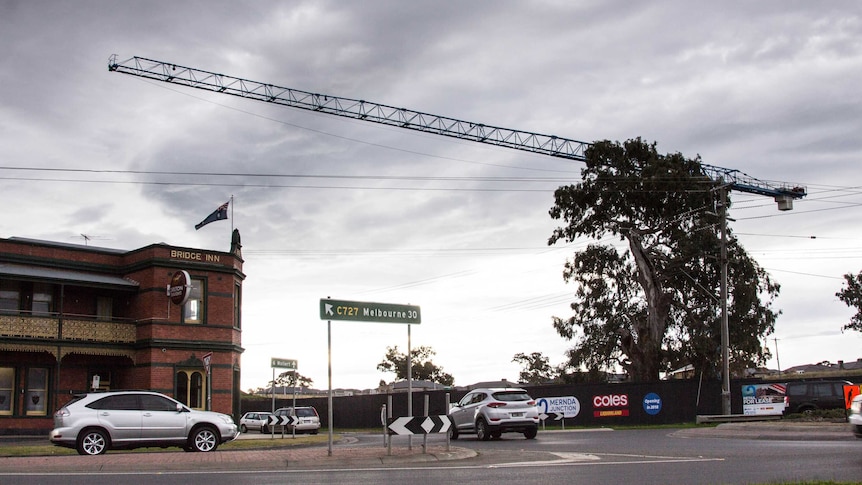 A large crane hangs over a busy intersection, country pub on one corner, new houses in background.