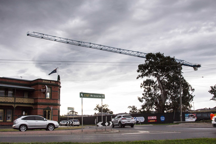 A large crane hangs over a busy intersection, country pub on one corner, new houses in background.
