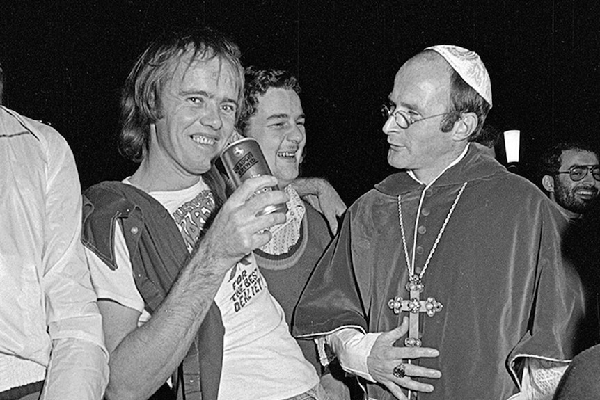 A man smiles in a crowd, holding a beer, next to another man dressed as a pope