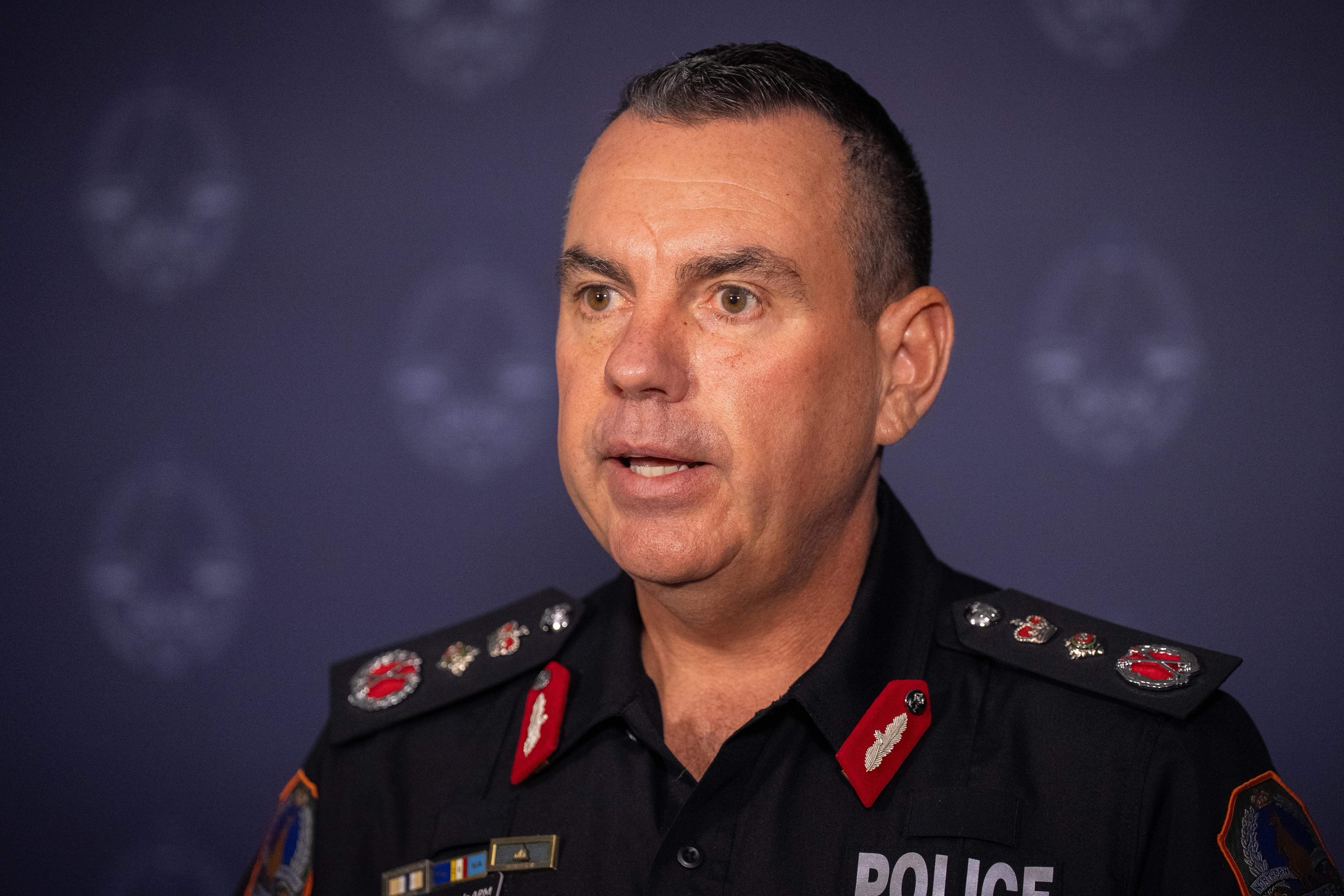 Close up of white man, with black short cut hair in a black police uniform, with a purple branded police banner behind him.