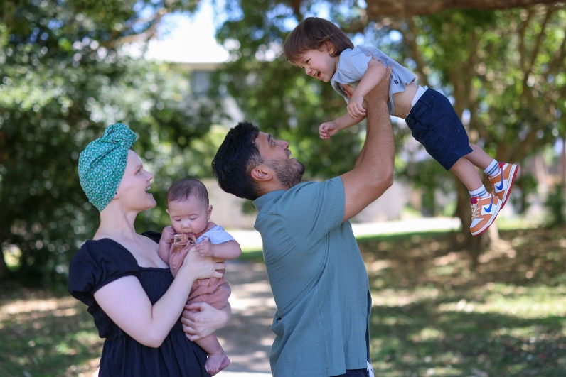 a man swings his toddler in the air as a woman in a headscarf holding a baby looks on