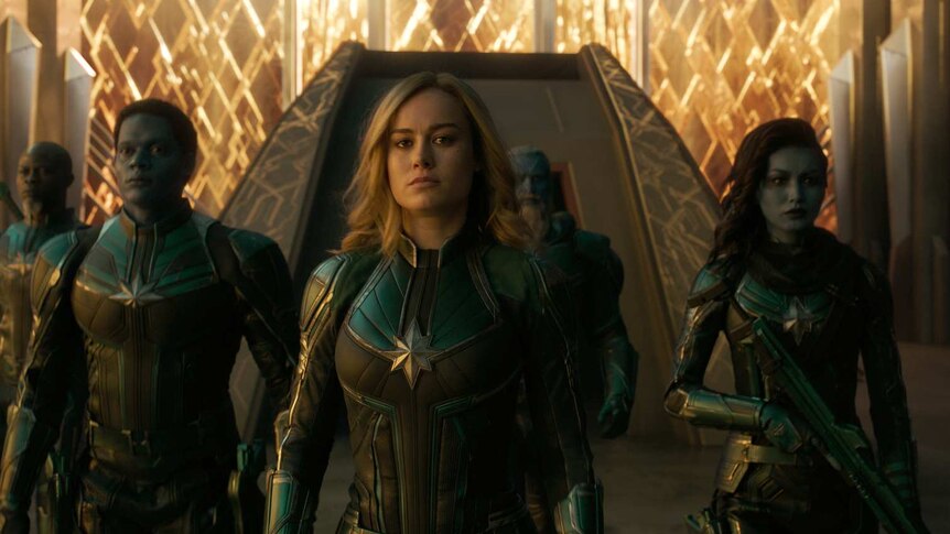 Larson and cast stand in formation, in green and black superhero costumes against a futuristic structure.