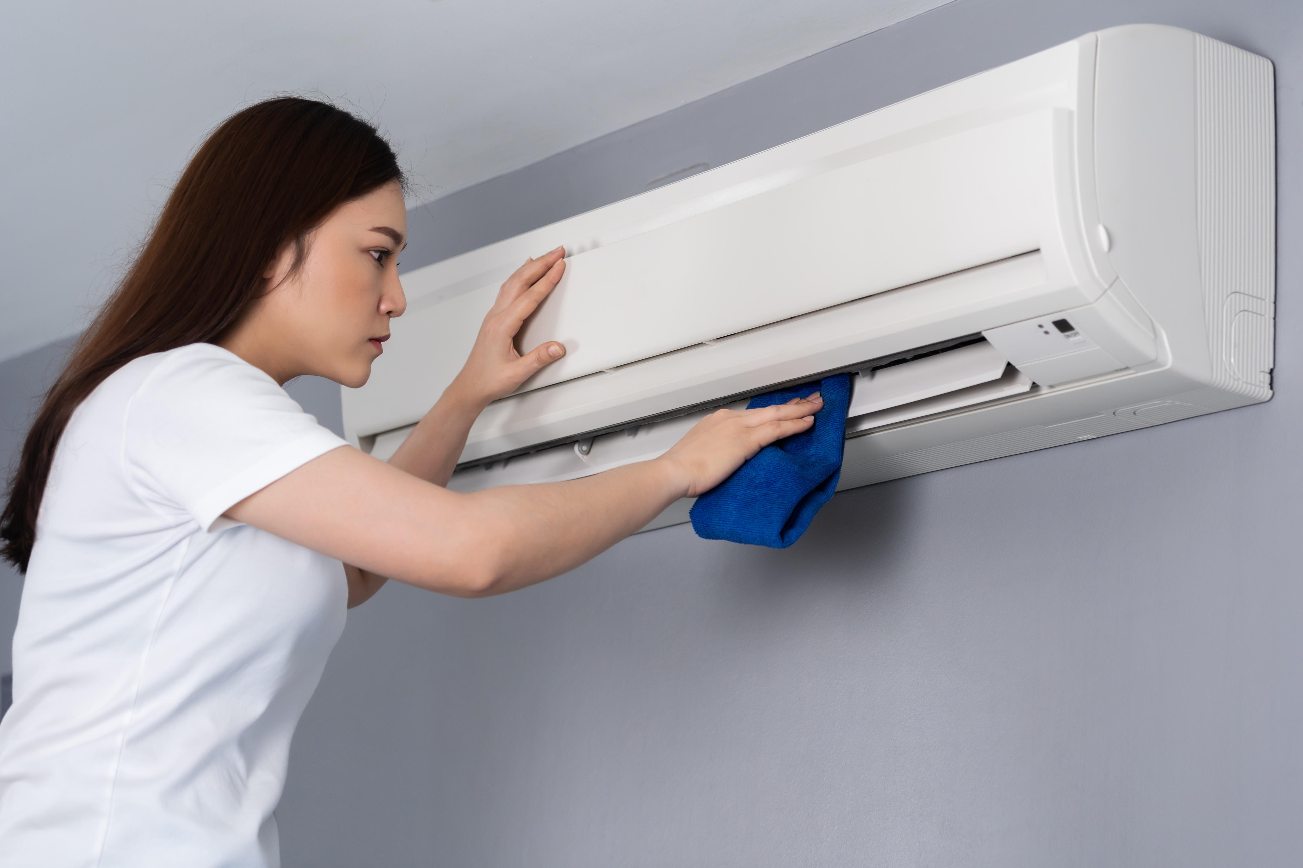 A woman of asian appearance with long brown hair wipes an air conditioner with a soft blue cloth