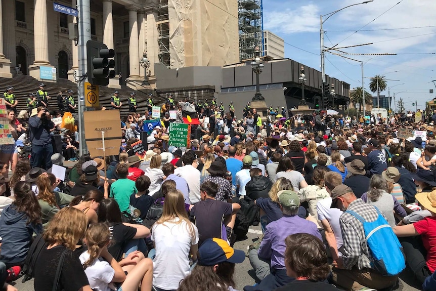 A large crowd of people sit in front of Victoria's Parliament House.