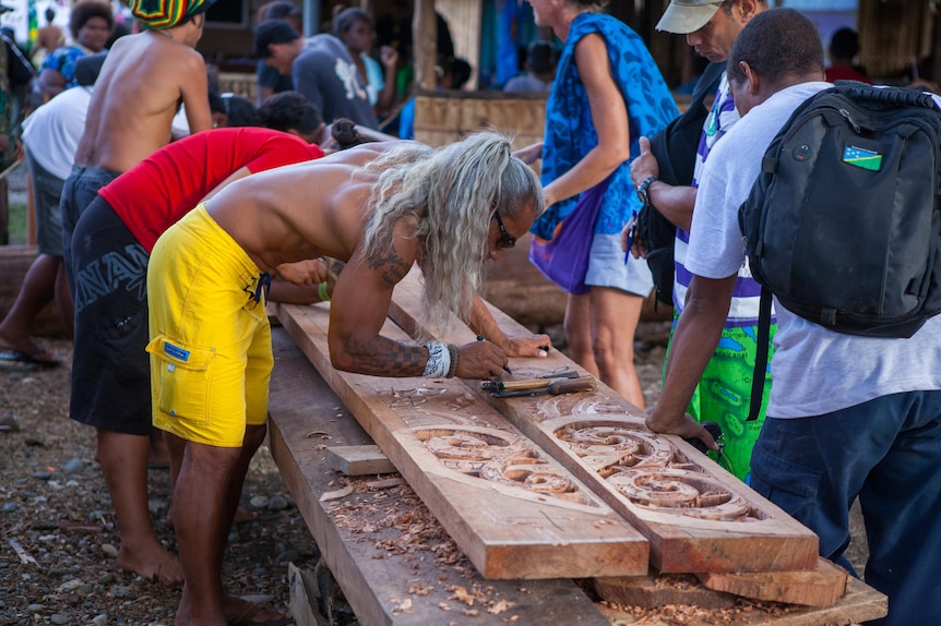 Man in yellow shorts and long grey hair stands over a large wood plank carving, surrounded by other carvers and onlookers. 