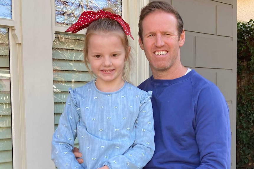 A young girl with a red bow in her hair sits on her father's lap outside their home.