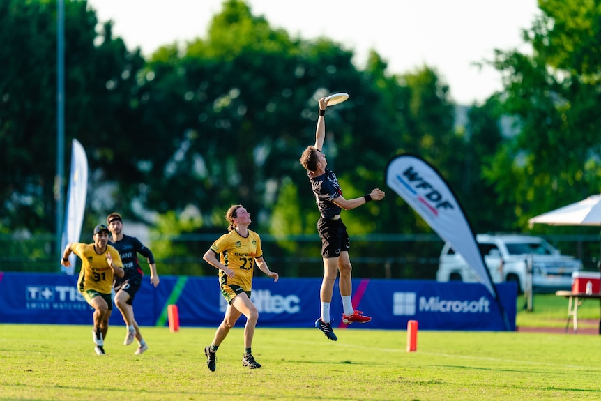 A man wearing dark colours jumps into the sky to catch a flying disc during a game against a team wearing yellow and green