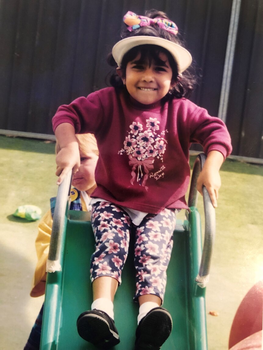 Tahnee at three years old sitting on a slippery slide wearing a had and maroon coloured jumper with floral pants.