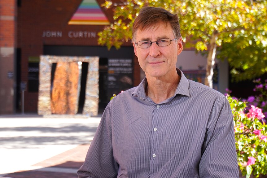 A man wearing a button up shirt and glasses, sitting outside with trees and the John Curtin centre in the background.