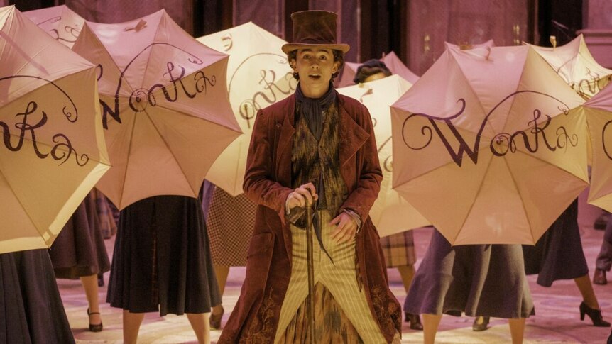Timothee Chalamet dressed as Wonka in a film still with people holding umbrellas saying WONKA