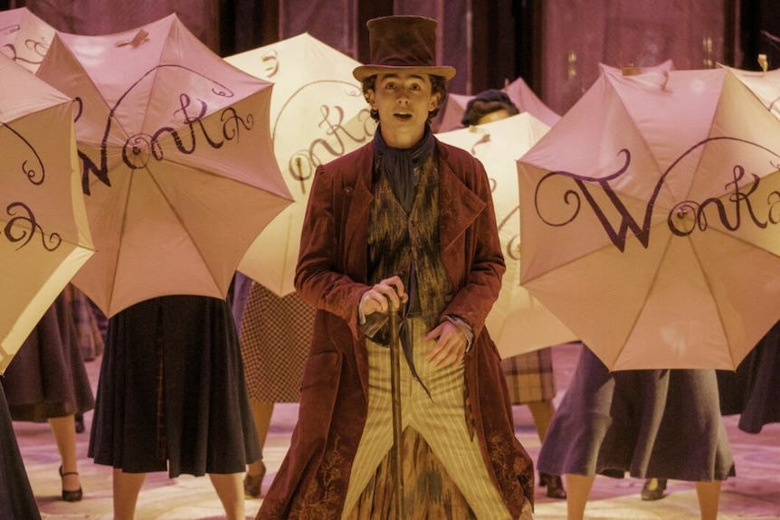 Timothee Chalamet dressed as Wonka in a film still with people holding umbrellas saying WONKA