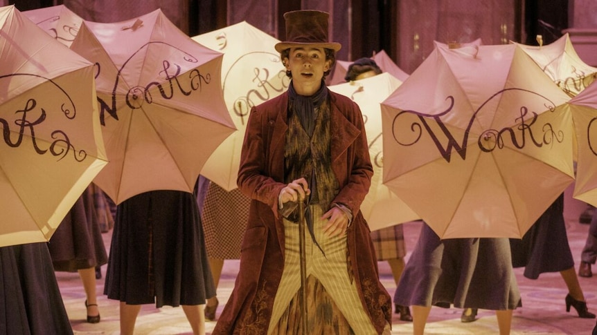 Timothee Chalamet dressed as Wonka in a film still with people holding umbrellas saying WONKA