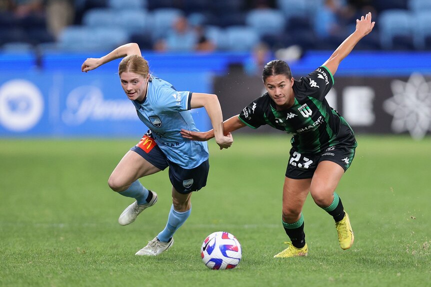 Two soccer players, one wearing blue and one wearing green and black, run for a ball during a match