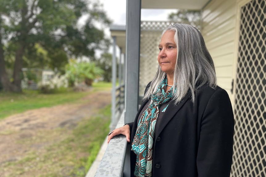 A woman with long grey hair in a black jacket stands outside a cabin.