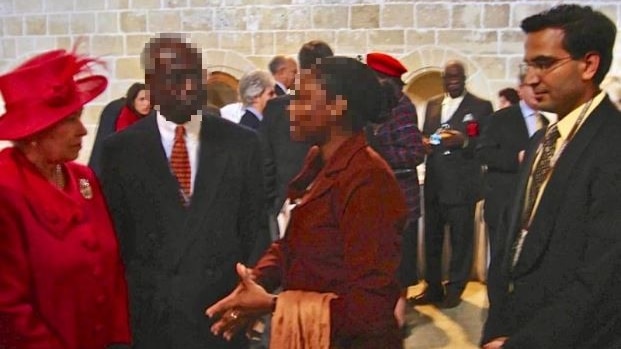 Queen Elizabeth stands to the left and a tall man in a black suit stands to the right, with blurred people in the middle. 